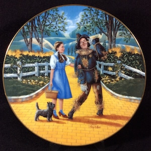 Puede incluir: Un plato de cerámica con una ilustración colorida de Dorothy de El Mago de Oz, el Espantapájaros y Toto caminando por el camino de baldosas amarillas. El plato tiene un borde dorado y un fondo azul.