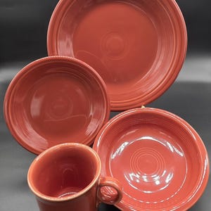 May include: A set of terracotta-colored ceramic dishware, including a large plate, a medium plate, a bowl, a smaller bowl, and a mug with a handle. The pieces have a glossy finish and concentric circle detailing.