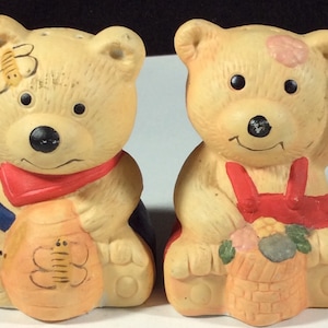May include: A set of two ceramic teddy bear salt and pepper shakers. The bear on the left is holding a honey pot with a bee on it. The bear on the right is holding a basket of flowers.