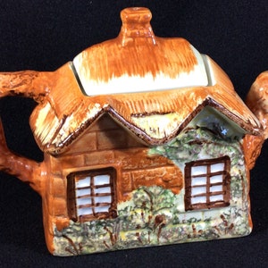 Puede incluir: Una tetera de cerámica marrón y blanca con forma de cabaña con techo de paja. La tetera tiene un asa y un pico marrones y dos ventanas con cristales blancos. La tetera está decorada con detalles verdes y marrones.