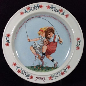May include: A white ceramic plate with a pink floral border. The center of the plate features a painting of two girls skipping rope. The text "A certified plate in the original and only Sarah Stilwell Weber Calendar Collection." is printed around the painting. The month "JUNE" is printed at the bottom of the plate.