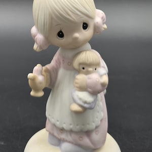 May include: A porcelain figurine of a child with blonde hair tied in a bow, wearing a pink dress with a white apron featuring a heart pattern. The child holds a small doll in one arm and a small object in the other hand, standing on a round base.
