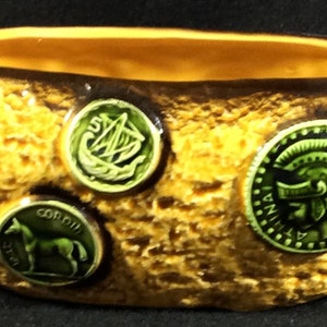 May include: A small, oval-shaped ceramic dish with a textured, yellow-brown exterior. The rim is dark brown, and the interior is a lighter yellow. Three green coin-like embellishments are affixed to the exterior, each with a different design.
