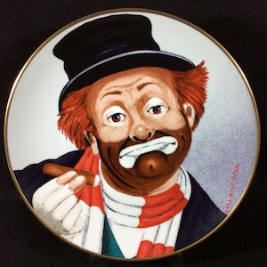 May include: A porcelain plate featuring a painting of a sad clown with red hair, a black top hat, and a red and white scarf. The clown is holding a cigar in his right hand. The plate has a gold rim and the signature "Jeff Vanuga 1989" is visible in the lower right corner.