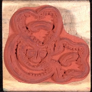 May include: A red rubber stamp with a detailed design of a stylized heart shape. The stamp is mounted on a wooden block.