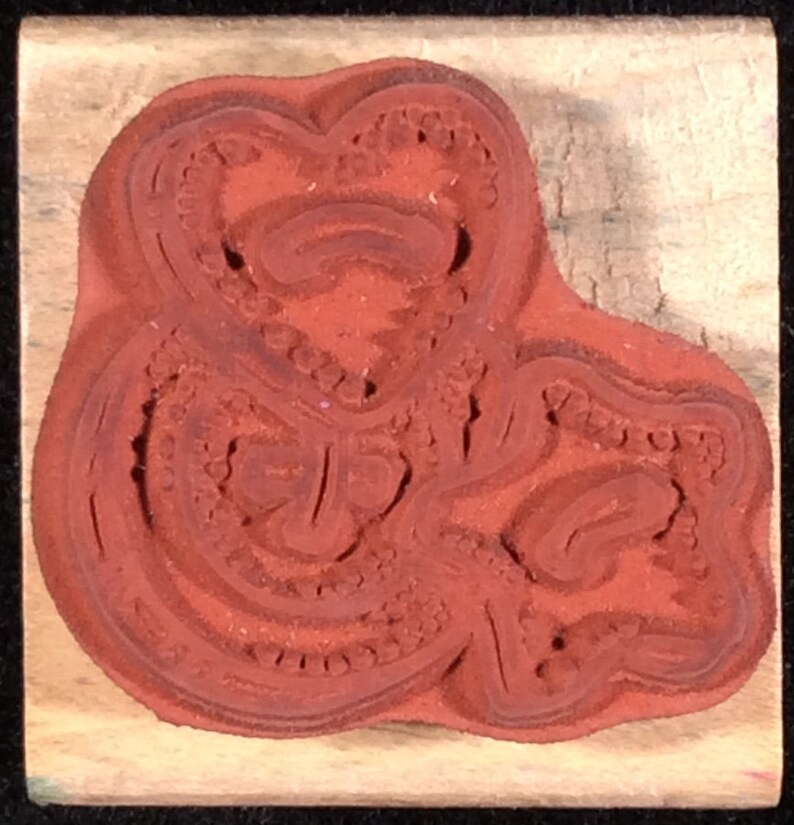 May include: A red rubber stamp with a detailed design of a stylized heart shape. The stamp is mounted on a wooden block.