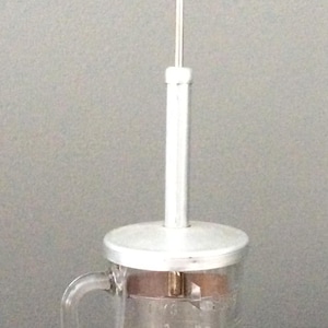 May include: A silver and glass kitchen tool with a red knob on top. The tool has a glass jar with a handle and a metal plunger that goes up and down. The jar has markings for measuring.