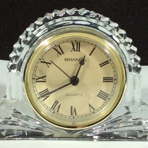 Puede incluir: Un reloj de cristal con una esfera dorada y números romanos. La esfera del reloj dice "Shannon Quartz".