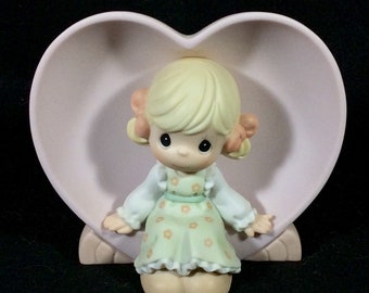 Puede incluir: Una figurita de porcelana de una niña con cabello rubio y un vestido verde sentada en un marco de corazón rosa.