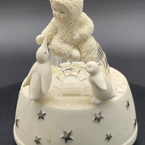 May include: A decorative figurine featuring a baby in a snowsuit sitting atop a clear igloo. Two white penguins stand on either side. The base is cream-colored with silver star accents, creating a winter theme.