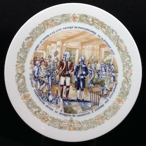 Puede incluir: Un plato de cerámica blanca con una ilustración colorida que representa una escena histórica. La escena muestra a George Washington invitando al marqués de Lafayette a unirse a su estado mayor después de una cena en la City Tavern de Filadelfia el 31 de julio de 1777. El texto en el plato dice "APRES UN DINER A LA CITY TAVERN DE PHILADELPHIE LE 31 JUILLET 1777 GEORGE WASHINGTON INVITE LE MARQUIS DE LAFAYETTE A JOINDRE SON ETAT MAJOR."