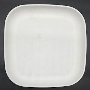 Puede incluir: Un plato cuadrado de cerámica blanca con esquinas redondeadas. El plato tiene una superficie lisa y está sobre un fondo oscuro. El plato está vacío y listo para la comida.