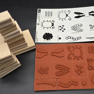 May include: A collection of rubber stamps with wooden blocks. The stamps feature various designs, including insects, clouds, and abstract shapes. The stamps are on a red rubber base and a white sheet. The wooden blocks are stacked.