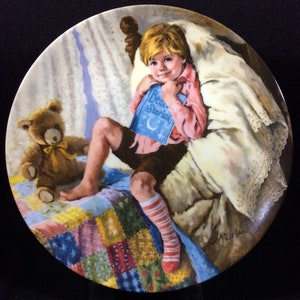 May include: A porcelain plate with a painting of a young boy in bed reading a book titled "Bedtime Stories". He is wearing a pink shirt, brown shorts, and striped socks. A teddy bear sits next to him on a colorful patchwork quilt.