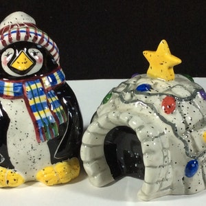 May include: A ceramic penguin figurine wearing a red and white striped hat and a blue and yellow scarf, standing next to a ceramic igloo figurine decorated with a yellow star and colorful lights.