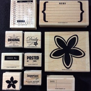 May include: A set of ten wooden rubber stamps with various designs and text. The stamps include a calendar, a memo, a flower, a note, a date, a flower, a note, a flower, a note, and a note. The stamps are all in a light wood color with black ink.