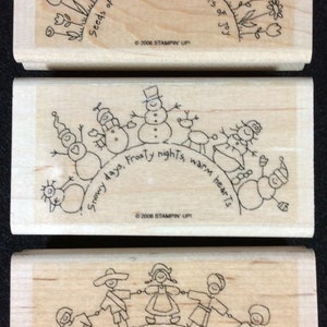 May include: Three wooden stamps with black ink illustrations and text. The top stamp features a semicircle of flowers with the text "Seeds of Friendship Bring Flowers of Joy". The middle stamp features a semicircle of snowmen with the text "Snowy days, Frosty nights, warm hearts". The bottom stamp features a semicircle of people holding hands with the text "Everyone smiles in the same language".
