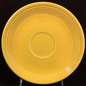 May include: A yellow ceramic saucer with a raised rim and a center well. The saucer has a glossy finish.