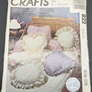 May include: McCall's Crafts pattern envelope showing different pillow designs. The pillows are in heart, round, and square shapes with frilled edges. The color scheme includes soft pinks, creams, and lavender. The text on the envelope reads "Pillow Essentials".