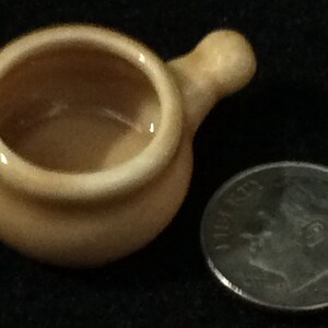 May include: A small, beige ceramic pot with a handle. The pot is about the size of a dime.