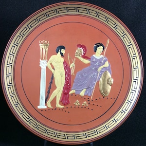 Puede incluir: Un plato de cerámica rojo con un borde dorado en forma de clave griega. El plato presenta una escena pintada de dos figuras, un hombre y una mujer, en estilo clásico. El hombre está desnudo y sostiene una lanza, mientras que la mujer lleva una túnica morada y sostiene un escudo. La escena se desarrolla frente a una columna blanca con una parte superior dorada.