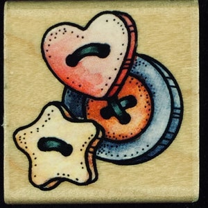 May include: Three fabric buttons in the shape of a heart, a star, and a circle. The heart button is pink and red, the star button is yellow, and the circle button is orange with a blue rim.