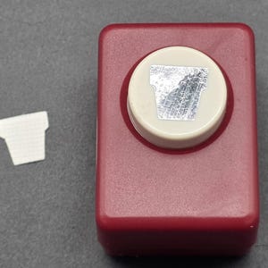 May include: A red and cream-colored paper punch with a flower pot design. A small, white paper flower pot cutout is next to the punch. The punch is made of plastic and has a lever for easy use. Ideal for scrapbooking and crafting.