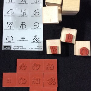 May include: A set of 12 wooden alphabet number stamps in a contemporary style. The stamps are arranged in a grid pattern on a white background. The stamps are made of wood and have a red rubber surface. The stamps are labeled with the numbers 1 through 9, 0, and 99. The stamps are also labeled with the ampersand symbol. The stamps are packaged in a clear plastic bag. ©2004 Stampin' Up! Contemporary Alphabet Numbers Set of 12