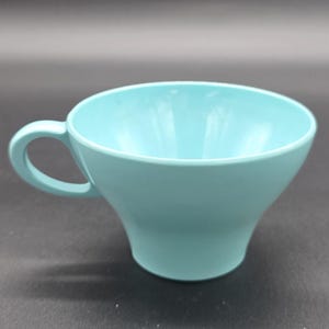 May include: A light blue teacup with a curved handle. The cup has a flared shape, wider at the top and narrowing towards the base. The cup is made of a solid, opaque material.