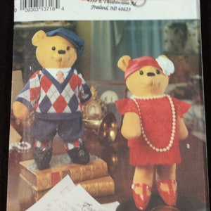 May include: Simplicity pattern 8263 for making two teddy bears. One bear is wearing a red dress with a white flower and a pearl necklace. The other bear is wearing a blue and red checkered sweater, blue pants, and a blue hat. The pattern is for crafts.