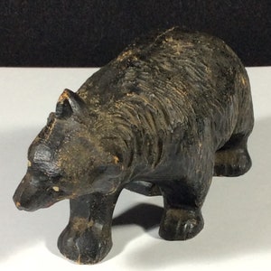 May include: A black wooden carved bear figurine. The bear is standing on all fours and has a detailed face with a small nose and eyes. The bear is in a walking position with its head turned to the side.