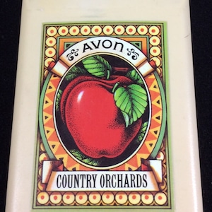Puede incluir: Un compacto vintage Avon Country Orchards con una manzana roja y hojas verdes en un marco decorativo. El compacto es de color crema con un borde dorado.