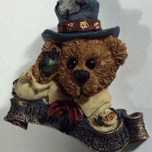 May include: A brown teddy bear wearing a blue top hat with a gold band and a red bow tie. The bear is holding a green object in its paw. The bear is sitting on a scroll with the text "The Original 1996-97".