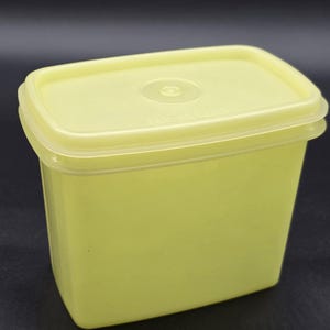 Puede incluir: Un recipiente rectangular amarillo pálido para guardar alimentos con una tapa a juego. La tapa tiene un centro ligeramente elevado con una hendidura circular. El recipiente está hecho de un material plástico duradero, adecuado para la cocina.