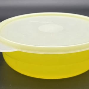 Puede incluir: Contenedor redondo amarillo para guardar alimentos con tapa a juego. La tapa es blanquecina y tiene una pequeña hendidura circular en el centro. Una pestaña blanca en forma de pétalo se extiende desde el lateral de la tapa. El contenedor es probablemente de plástico.