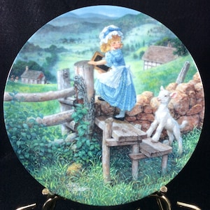 May include: A blue and white porcelain plate with a painting of a girl in a blue dress and a white hat holding a book. The girl is standing on a wooden fence and looking at a white lamb standing on a stone wall. The background is a green field with a small house in the distance.
