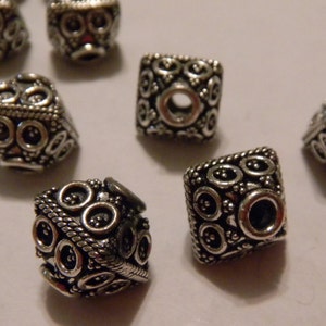 May include: A group of eight silver-toned square beads with intricate designs. The beads have a hole in the center for stringing.
