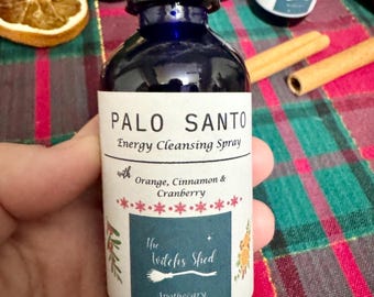 Peut inclure: Une bouteille en verre bleu avec une étiquette blanche qui indique "Palo Santo Energy Cleansing Spray with Orange, Cinnamon & Cranberry The Witches Stied Apothecary".