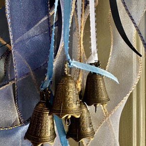 Op de afbeelding: Close-up van vier kleine, bronskleurige belletjes die aan blauwe, witte en beige linten hangen. De belletjes hebben ingewikkelde gegraveerde patronen en hangen tegen een achtergrond van gelaagde, transparante linten in blauw- en wittinten.
