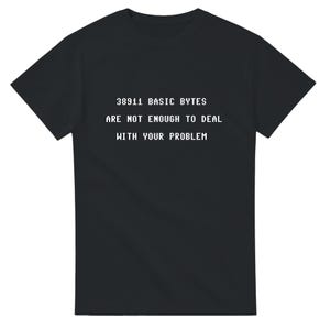 Puede incluir: Camiseta negra con texto blanco: "38911 BASIC BYTES ARE NOT ENOUGH TO DEAL WITH YOUR PROBLEM". El texto está en una fuente retro pixelada. La camiseta es de cuello redondo.