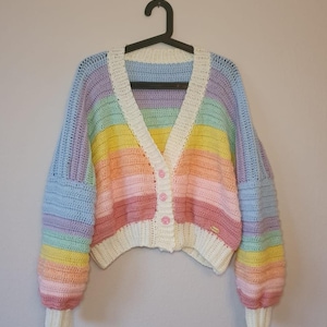 May include: A pastel rainbow-striped crochet cardigan with a V-neck and long sleeves. The colours include blue, purple, yellow, pink, and white. The cardigan has a white trim and is fastened with pink buttons. The sleeves have a ribbed cuff.