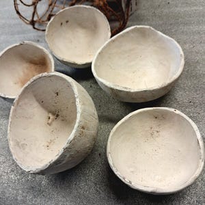 Könnte beinhalten: Fünf cremefarbene, handgefertigte Schalen mit strukturierter Oberfläche sind auf einer grauen Oberfläche angeordnet. Im Hintergrund ist ein geflochtener Korb zu sehen. Die Schalen haben eine rustikale, natürliche Ästhetik.
