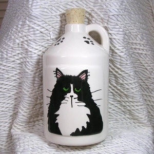 May include: White ceramic jug with a black and white cat illustration. The cat has green eyes and a white patch on its chest. The jug has a cork stopper and a handle.