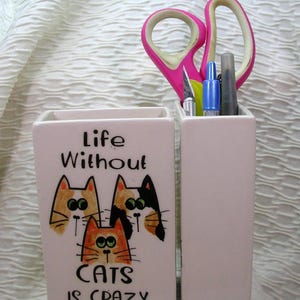 May include: A two-part ceramic desk organizer with a light pink finish. One section features the text "Life Without Cats Is Crazy" and cat illustrations. The other section holds pink scissors, a blue pen, and other tools.