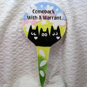 May include: A whimsical garden stake with a round top featuring the text "Comeback With A Warrant." Below the text are three black cat silhouettes with heart and eye details. The stake has a colorful sky and grass design.