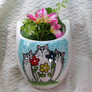 May include: White ceramic wall vase with a whimsical cat and flower design. The vase features three gray cats with flowers in red, yellow, and blue. Pink and white flowers and greenery are inside the vase.