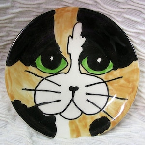 May include: A round ceramic dish featuring a hand-painted calico cat face. The design includes large green eyes, a white muzzle, black whiskers, and patches of black and tan fur against a white background, creating a detailed feline portrait.