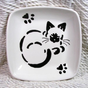 May include: A white ceramic dish with a black silhouette of a cat curled up sleeping. The cat has paw prints on either side of it.