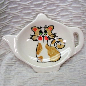 May include: White ceramic tea bag holder in the shape of a teapot, featuring a hand-painted cartoon cat with brown and white spots, green eyes, and red cheeks. The holder has a spout and a handle.