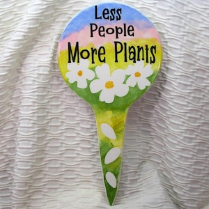 May include: A garden stake with a colorful design. The top is a circle with the text "Less People More Plants" in black. White daisies with yellow centers are painted below the text. The stake has a green and yellow background.